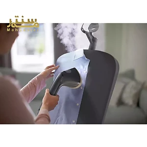 در تصویر، یک خانم در حال استفاده از دستگاه بخارگر برای صاف کردن چروک‌های یک پیراهن مردانه آبی رنگ دیده می‌شود.