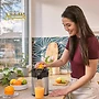 یک زن در آشپزخانه‌ای مدرن با لبخند در حال گرفتن آب پرتقال با یک آبمیوه‌گیری برقی است.