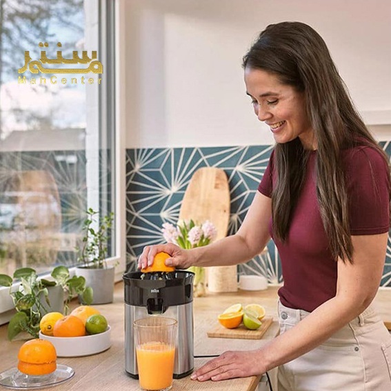 یک زن در آشپزخانه‌ای مدرن با لبخند در حال گرفتن آب پرتقال با یک آبمیوه‌گیری برقی است.