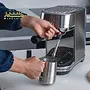 دستگاه اسپرسوساز استیل با طراحی مدرن، در حال تهیه قهوه، با کاربر در حال نصب فیلتر و استفاده از بخار برای تولید کف شیر، نشان‌دهنده فرآیند تهیه قهوه حرفه‌ای.