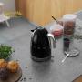 یک کتری مشکی SMEG در پس زمینه سفید نمایش داده می شود. کتری پر از آب است و روی پایه خود قرار دارد. دو فنجان چای در کنار کتری قرار دارند.