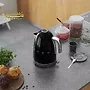یک کتری مشکی SMEG در پس زمینه سفید نمایش داده می شود. کتری پر از آب است و روی پایه خود قرار دارد. دو فنجان چای در کنار کتری قرار دارند.