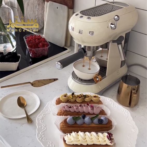 یک دستگاه اسپرسو کرم SMEG با فنجان های قهوه و نان های خامه ای در یک آشپزخانه نشان داده شده است.