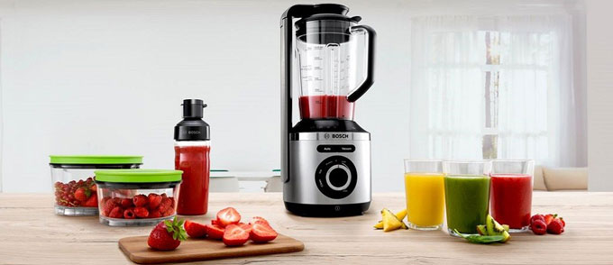 یک مخلوط کن bosch نقره‌ای و مشکی در کنار سه لیوان آبمیوه، دو ظرف میوه پر از توت فرنگی و یک بطری آبمیوه
